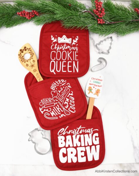Three red Christmas potholders stuffed with wooden spoons and cookie cutters. Add whimsical phrases like "Cookie Queen" and "Christmas Baking Crew" with your Cricut and Abbi's SVG files. 