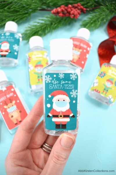 Abbi holds a "Santa-tizer" bottle with a Cricut sticker you can make as a last minute Christmas gift. Examples of other sanitizer and soap bottles lay in the background on a blue tabletop. 