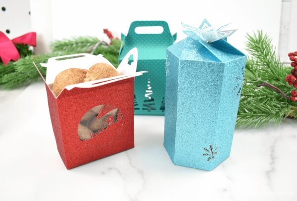 Red, blue and teal glitter foldable cookie boxes with a cellophane covered window in the sides. Christmas garland decorates the background. These cookie boxes make great Cricut last minute Christmas gifts. 