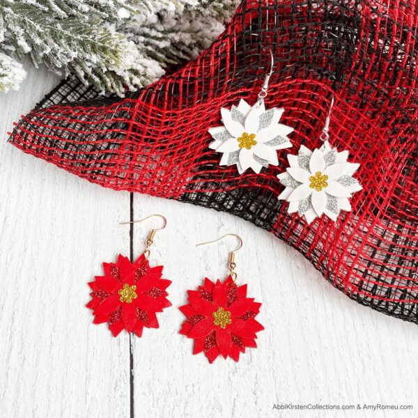 A pair of white Poinsettia Faux Leather Earrings hooked on red and black gauze fabric alongside a pair of red Poinsettia Faux Leather Earrings on a white table top.