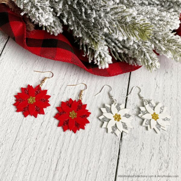 Two pairs of red and white Christmas Poinsettia Faux Leather Earrings on a wooden table next to a flocked pine branch. 