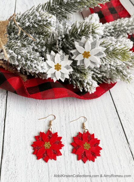 White Poinsettia Faux Leather Earrings hooked on a flocked pine branch alongside Red Poinsettia Faux Leather Earrings on a wooden white table. 