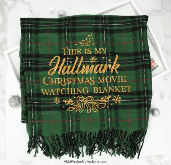 A green fleece blanket with gold iron-on vinyl that says "This is my Hallmark Christmas movie watching blanket." Make these blankets as last-minute DIY gifts with your Cricut. Get the free hallmark movie SVG on Abbi Kirsten Collections. 
