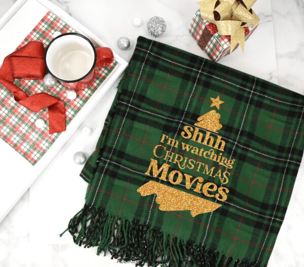 A plaid green fleece Christmas blanket with gold glitter iron-on vinyl that says "Shh I'm watching Christmas movies" surrounded by presents and bright red gift wraps. Get the free SVG file on Abbi Kirsten Collections. This is one of six Christmas gifts to make with Cricut.
