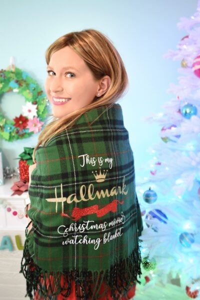 Abbi Kirsten models a plaid green blanket that says "This is my Hallmark Christmas movie watching blanket" in front of part of a white Christmas tree and a DIY paper wreath. This blanket makes a great Cricut last minute gift. 