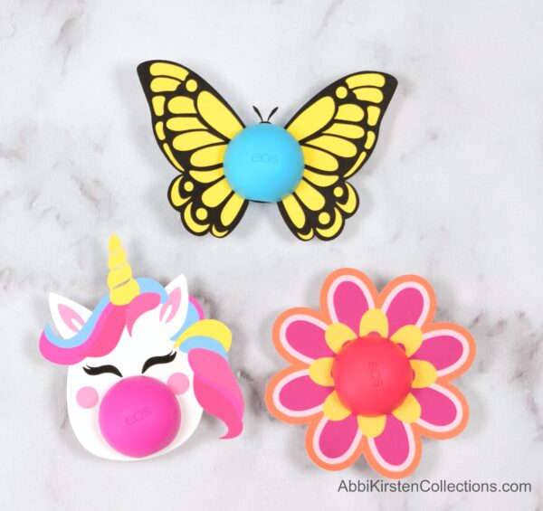 A close-up overhead view of the three free printables for Eos lip balms include a unicorn, a pink flower, and a yellow butterfly. 