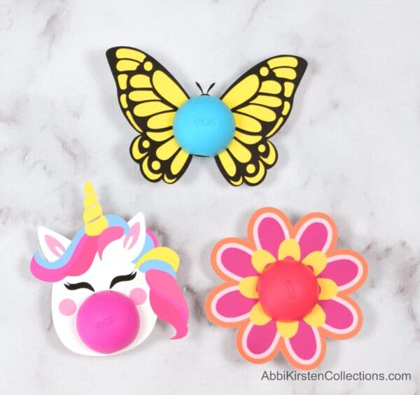 A close-up overhead view of the three free printables for Eos lip balms include a unicorn, a pink flower, and a yellow butterfly. 