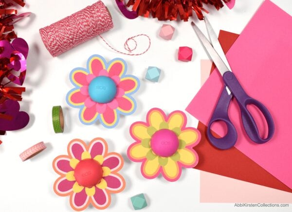 Free flower printables use the Eos lip balms as the flower center. Scissors, pink paper, washi tape, and red and white string surround the decorated lip balm.