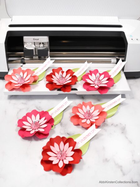 Six red and light pink Paper flower Kiss candy valentines next to and on top of a Cricut machine.