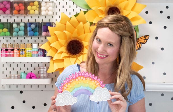 Abbi smiles standing in her craft room, holding the layered 3D rainbow mandala paper craft made with a Cricut machine. 