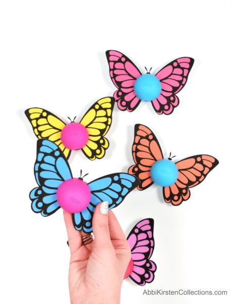 Abbi Kirsten's hand holds a butterfly Eos printable, above other colorful butterflies laying on a white table. 