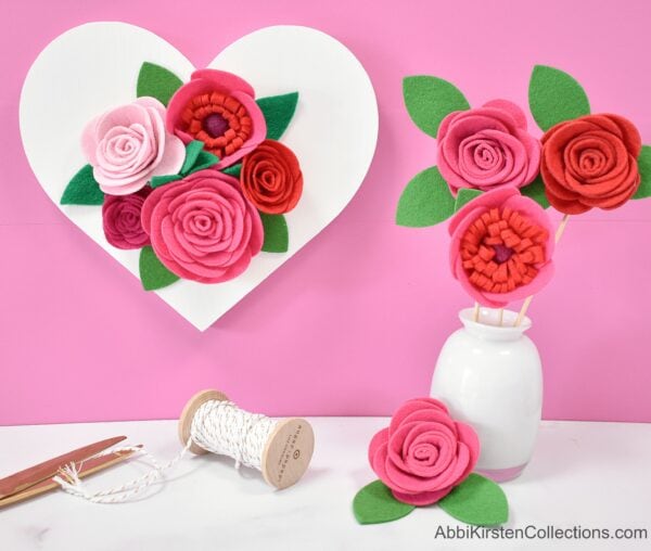 Felt roses in pink and red decorate the pink wall, the white table, and sit in a white vase. A roll of twine and shiny scissors lay nearby. 