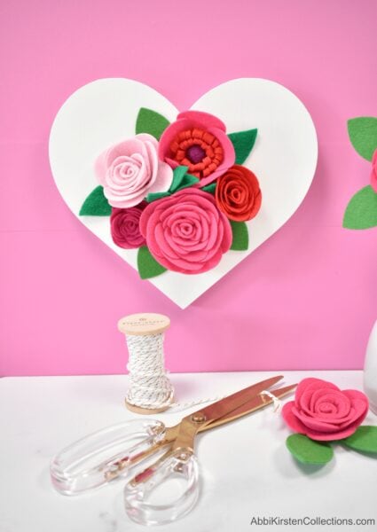 Scissors and twine lay on a white table with a finished felt rose. On the pink wall is a white wooden heart adorned with felt roses and greenery. You can use your Cricut or scissors for this felt craft. 