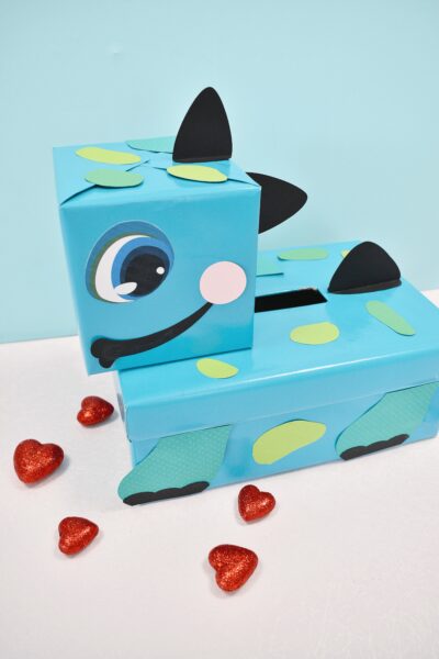 This adorable dinosaur Valentine box is perfect for kids' Valentine's Day at school! The light blue dinosaur box is dotted with green and yellow spots, black spikes, and a cute little smile.