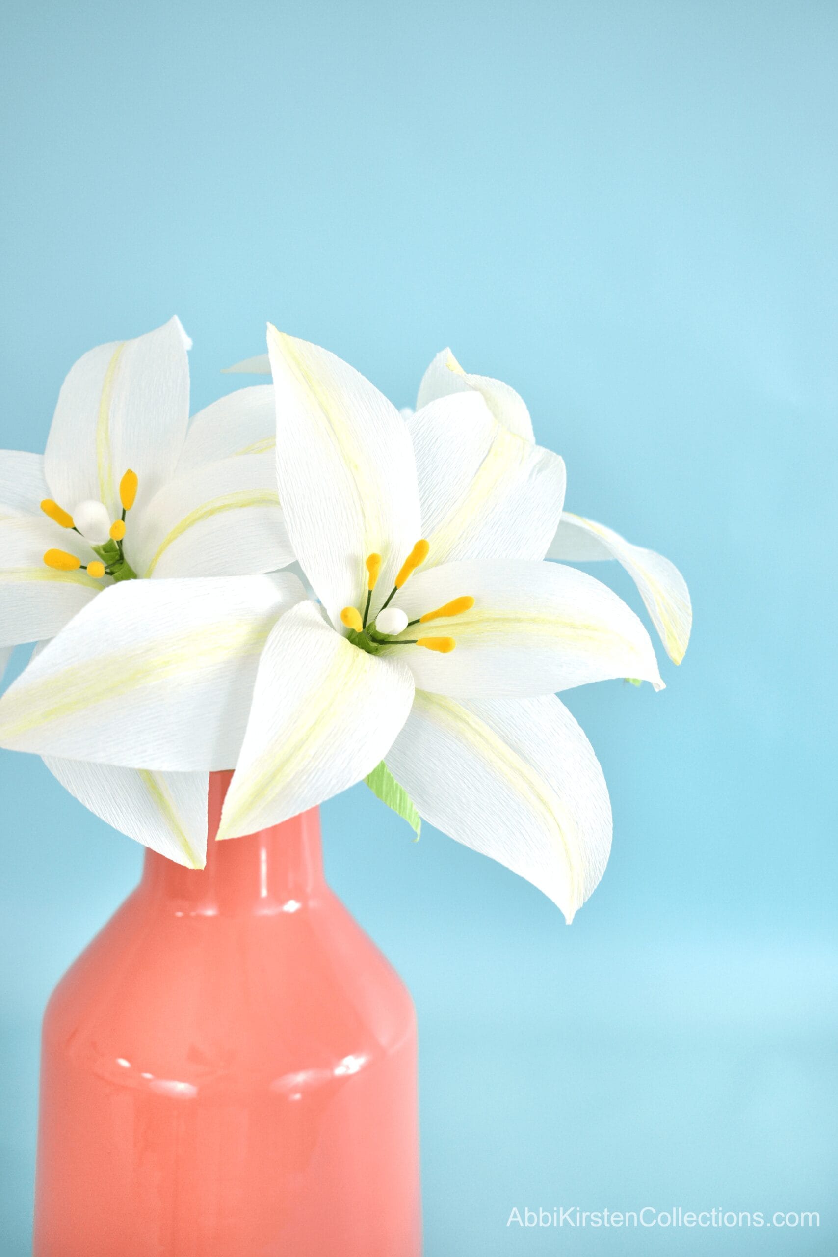 A bundle of crepe paper Easter lily flowers in a opaque salmon-colored ceramic vase in front of a light blue paper backdrop. 