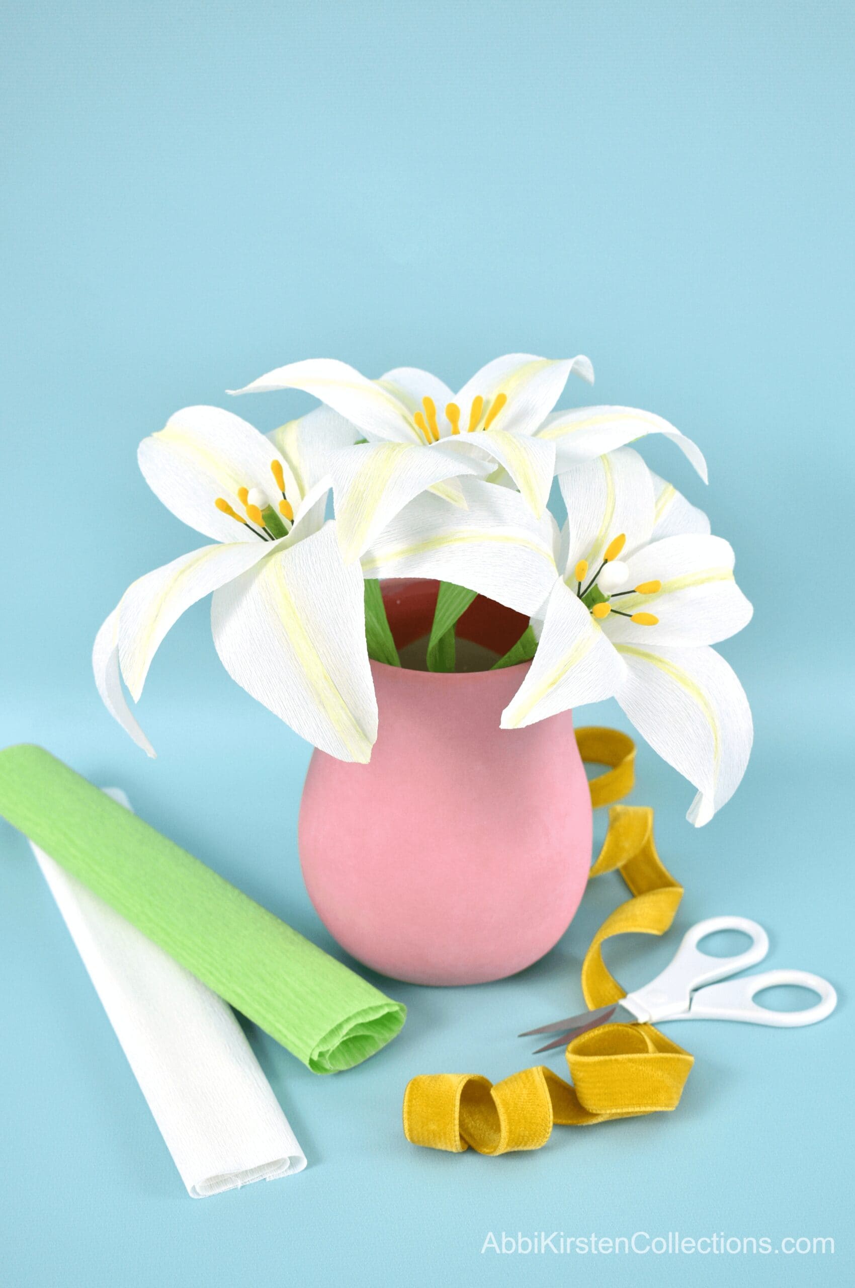 A finished bouquet of three white crepe paper lilies with green leaves in a pink vase. Rolls of white and green crepe paper, detail scissors and yellow ribbon lay in front of the vase on a light blus paper backdrop. 