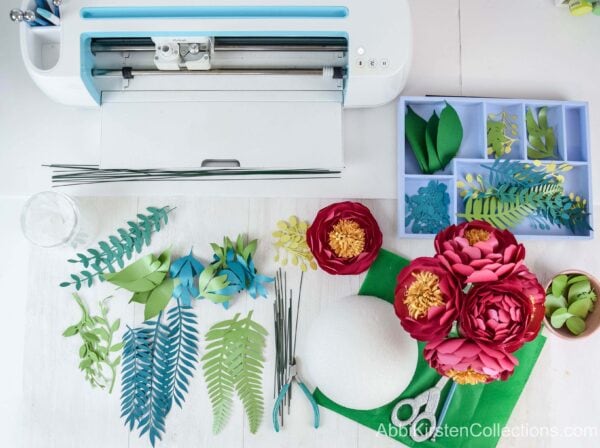 A bird’s eye view of the supplies needed to make a cascading paper flower bouquet, including pre-made flowers, greenery, and a Cricut machine.