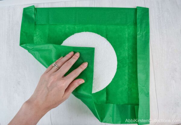 A square piece of green tissue paper lays under a foam base. A woman’s hand shows how to fold the tissue paper around the bouquet base. 