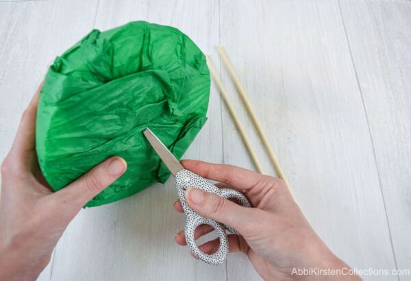 Hands holding crafting scissors poke a hole into a green tissue paper wrapped half foam ball at a 45-degree angle so that the bouquet handle can be inserted. 
