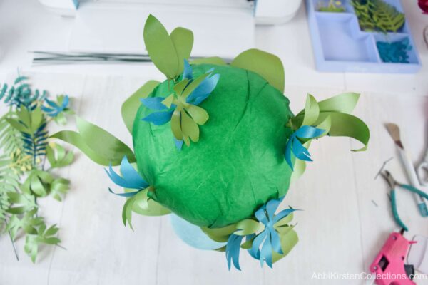 The green tissue paper-wrapped foam ball has various greenery added to it. It’s best to start with the larger paper leaves closer to the edges of the cascading bouquet. 