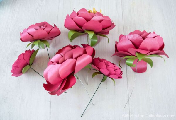 Six red paper peonies with wire stems and paper leaves lay on a workspace, ready to be added to a paper flower bouquet or arrangement. 