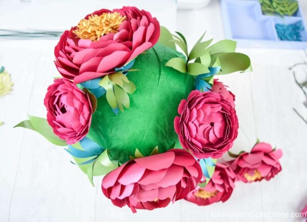 The foam base now has more extensive greenery, large and small pink paper peonies. The paper flower bouquet is built one flower at a time. 