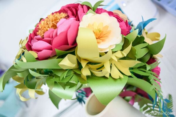 A top view of the cascading paper flower bouquet shows the half ball foam base shape, with lush greenery and yellow, white and red flowers blooming outward. 