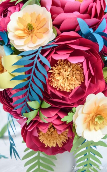 A close-up of the paper bouquet shows blue-green fern leaves, large and small red peonies, yellow and white buttercups, and blue and green leaves. The foam base is not visible. 