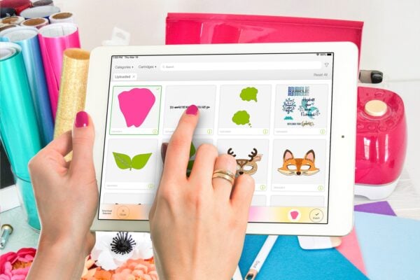 A woman's hands hold a white tablet, the screen displays the Cricut Design Space app.