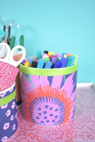 A cylinder vase filled with markers and decorated with pink, red and purple wrapping paper and green ribbon.