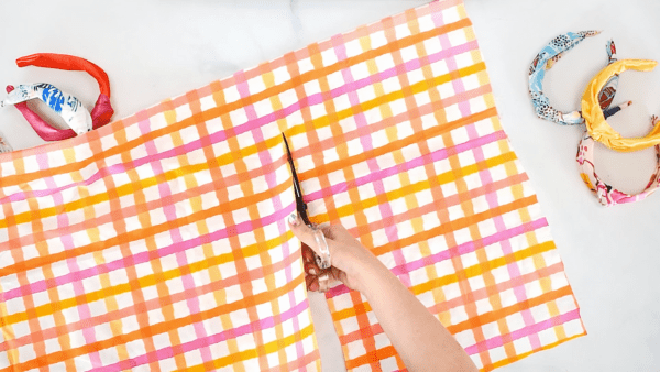Cutting a sheet of yellow, orange, and pink plaid fabric with scissors. If you don’t use a Cricut, you can cut the free top knot headband pattern yourself using fabric scissors.