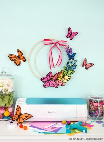 A paper butterfly wreath hangs on a light blue wall, surrounded by colorful paper butterflies fluttering around. Cricut supplies and cardstock are laid out on a table below.