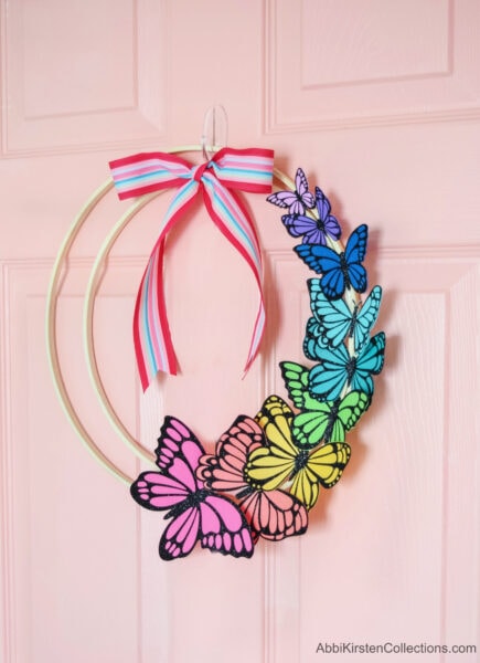 A DIY paper butterfly wreath hangs on a pink front door. It's decorated with a colorful red, blue and pink ribbon.