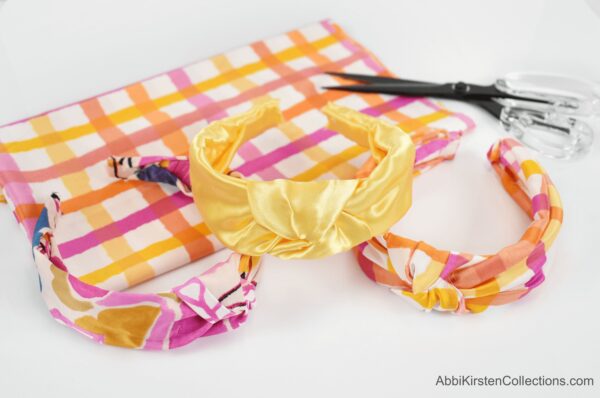 Three multicolored satin fabric headbands are displayed on a checkered piece of fabric. With this tutorial you can make a variety of no-sew top knot headbands. 