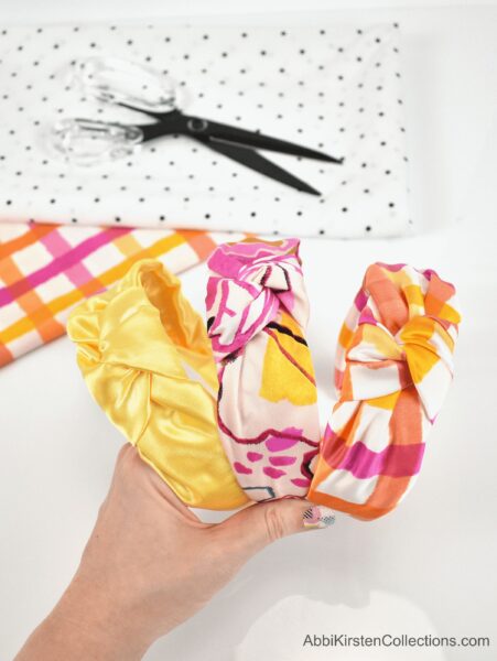 A woman’s hand holds yellow, orange, and pink DIY fabric no-sew headbands, fabric scissors, and a polka-dotted fabric sample for use in a baby headband. 