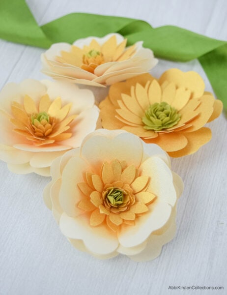 Three pale yellow and one bright yellow buttercup paper flowers next to a bright green ribbon on a white wooden table. 