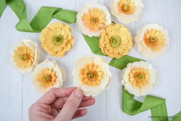 A bright green ribbon underneath nine pale yellow buttercup paper flowers with light green centers displayed on a white table.