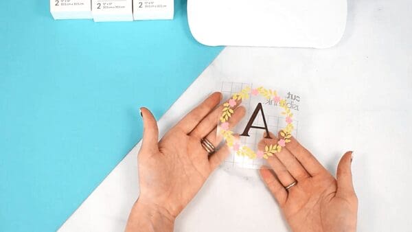 Abbi Kirsten holds a monogramed wreath design on transfer paper. Infusible ink boxes and a Cricut machine are partially seen from above. 