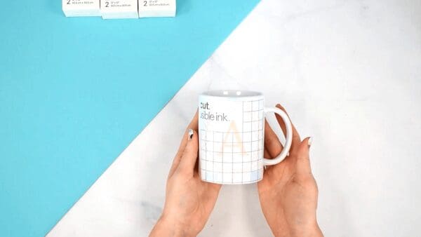 In her palms, Abbi Kirsten holds a white mug with transfer tape on the side. The design is made with infusible ink, which means the mug will be dishwasher safe. 