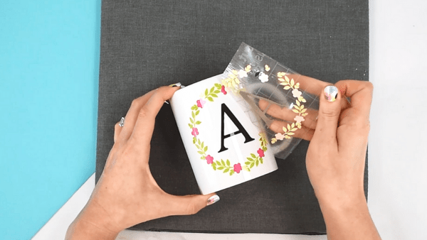 Abbi Kirsten's hands pull the transfer tape off the white mug revealing a  monogrammed letter "A" surrounded by a multi-colored wreath. 