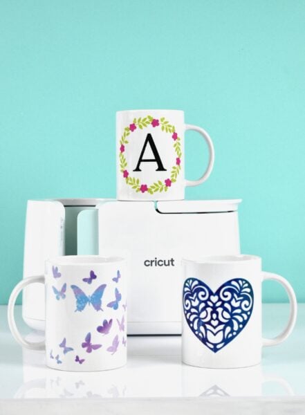 Three white mugs with carius designs on a table with a Cricut Mug Press. Learn how to use the Cricut mug press, and how you can add more than one color of infusible ink to your mug so that you can add different colors and layers to your design! 