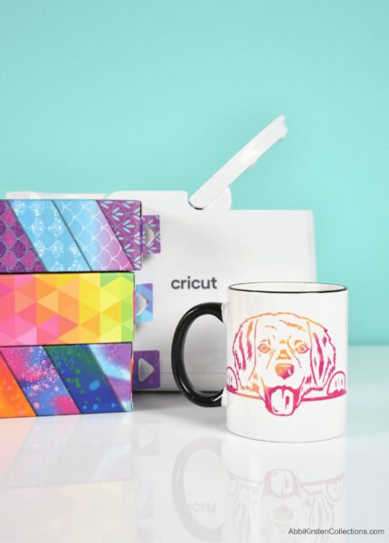 A Cricut Mug Press is behind boxes of infusible ink and a white mug with a rainbow dog design. 