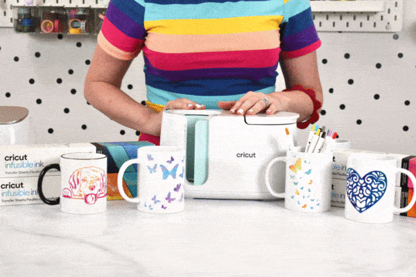 Abbi poses with a Cricut Mug Press, surrounded by boxes of infusible ink and mugs with unique designs. 