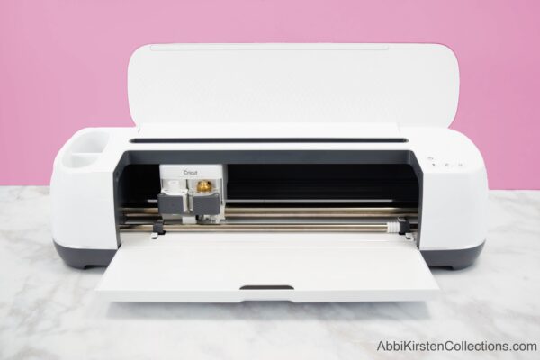 A white Cricut Explorer machine on a clean marble tabletop and a dusty rose background. This machine is easy to learn. 