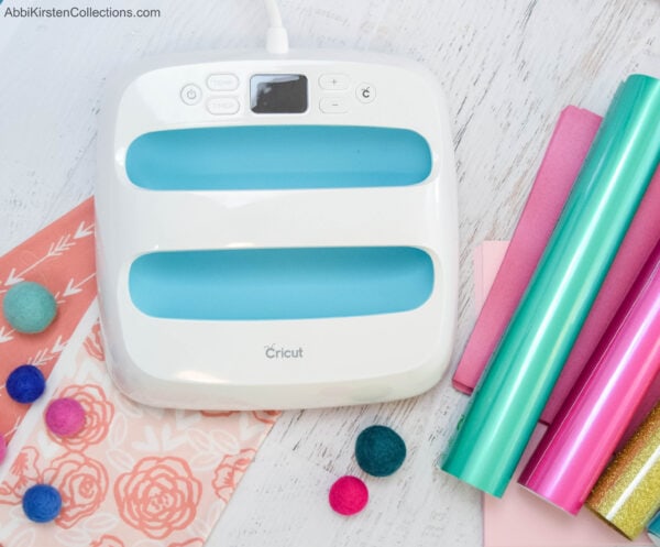 The Cricut Easypress is seen from above on a white wooden tabletop. Craft balls, rolls of candy-colored vinyl rolls, and other cut-outs are around the Easypress. 