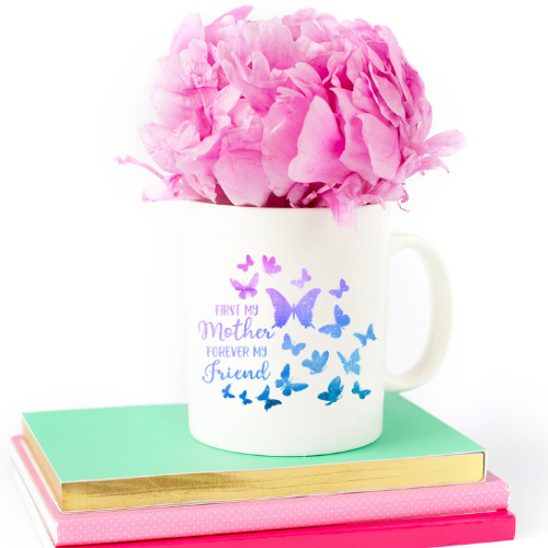 Pink crepe paper flower petals sit inside a white mug on top of colorful books. The mug has butterflies and the words "First My Mother, Forever My Friend."