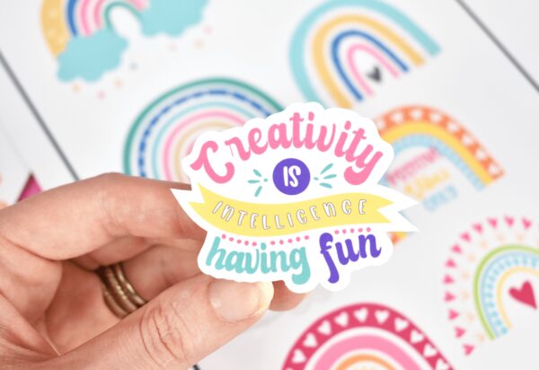 Abbi holds a sticker made with silky smooth printable vinyl. In pink, purple, green and yellow, the sticker says "creativity is intelligence having fun."