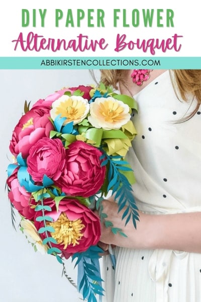 A woman holds a DIY cascading flower bouquet of red, green, white, and yellow paper flowers and paper leaves. 