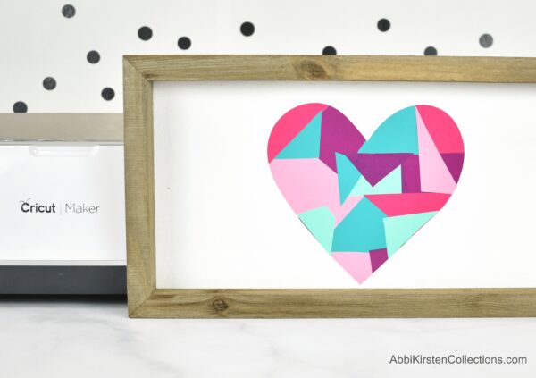A wooden frame with a white background and red, teal, pink and purple colored heart in the center. One Cricut vinyl hack is to create art using your vinyl scraps. 