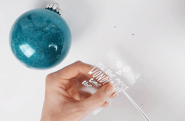 A woman's hand holds a piece of transfer tape with a Christmas message in vinyl. She is cutting the message so that it can be placed on the large blue green circular ornament next to here. More Cricut vinyl hacks to make crafting more fun. 